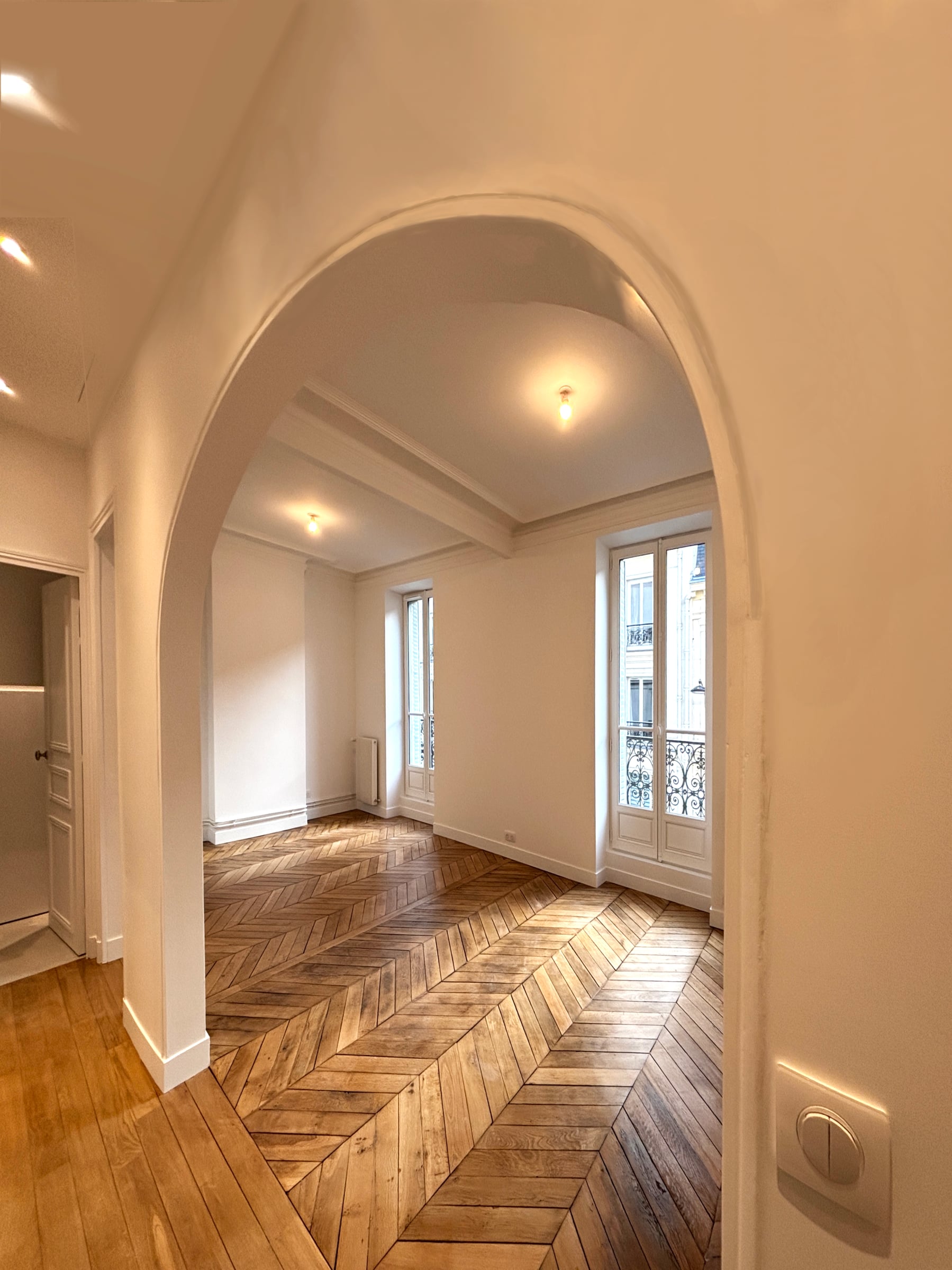 Entrée voûtée en plâtre ouvrant sur un séjour au parquet point de Hongrie, double porte-fenêtre à garde-corps en fer forgé.