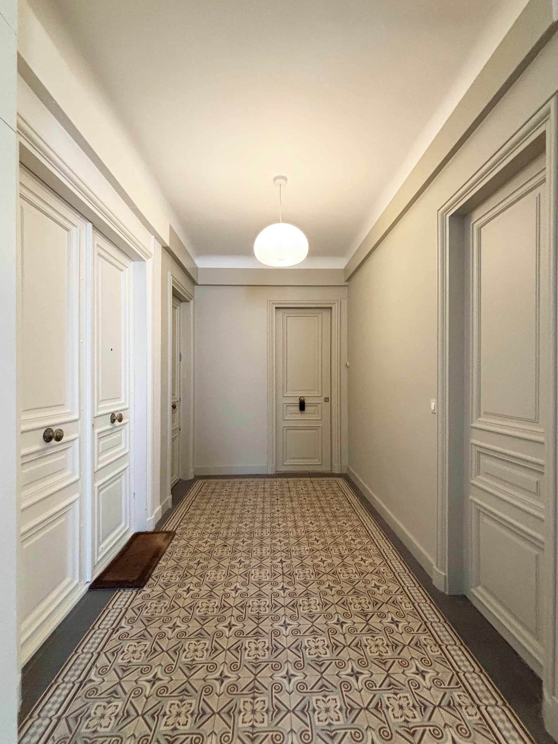 Restauration de couloir de copropriété avec portes à panneaux moulurés, sol à carreaux anciens et suspension blanche centrale.