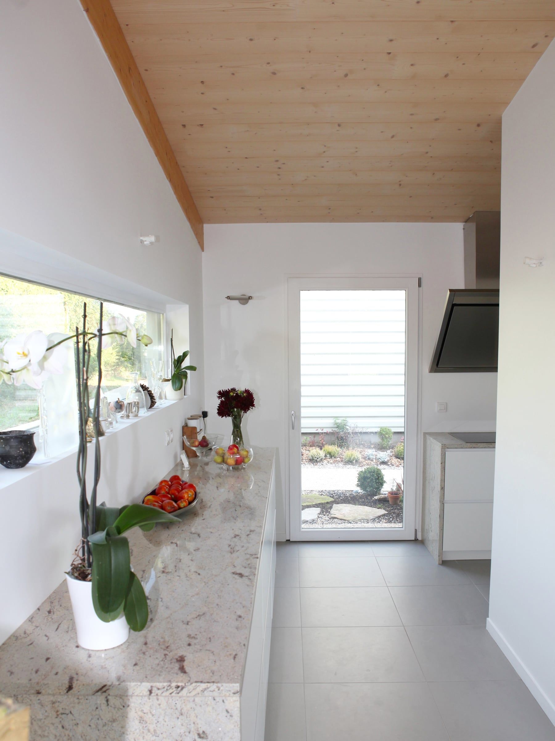 Rénovation de cuisine avec rangements blancs, plan de travail clair veiné, crédence-fenêtre sur le jardin et plafond lambrissé en bois.