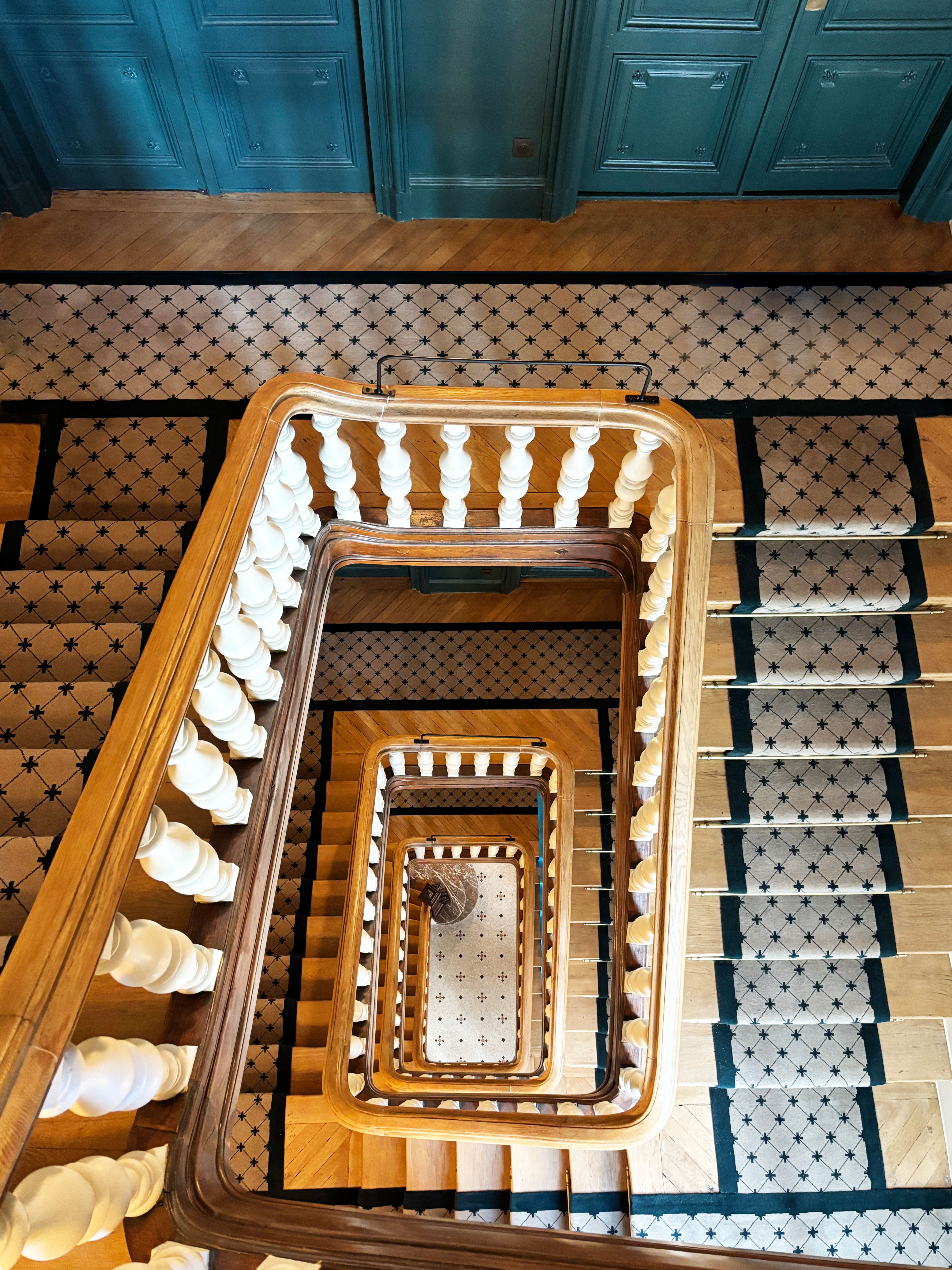 Plongée dans la cage d’escalier rénovée, mains courantes en bois, balustres blancs et tapis sur mesure à motifs sur parquet et mosaïque.
