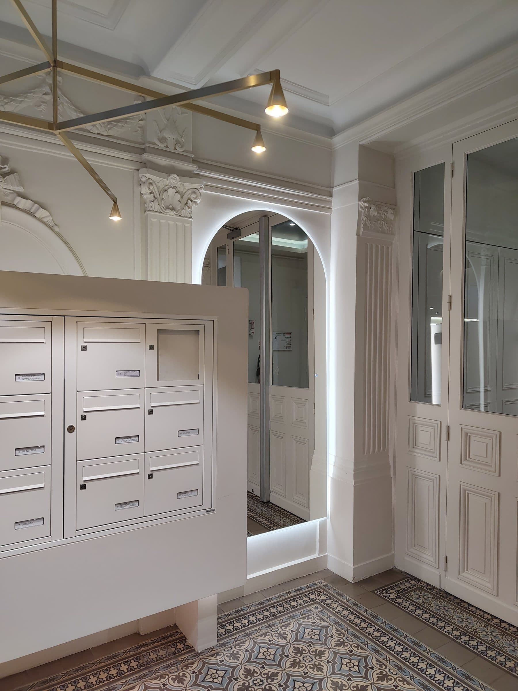 Après travaux : hall d'immeuble rénové sous haute verrière, ornements en plâtre restaurés, boîtes aux lettres encastrées et carreaux de ciment au mur.