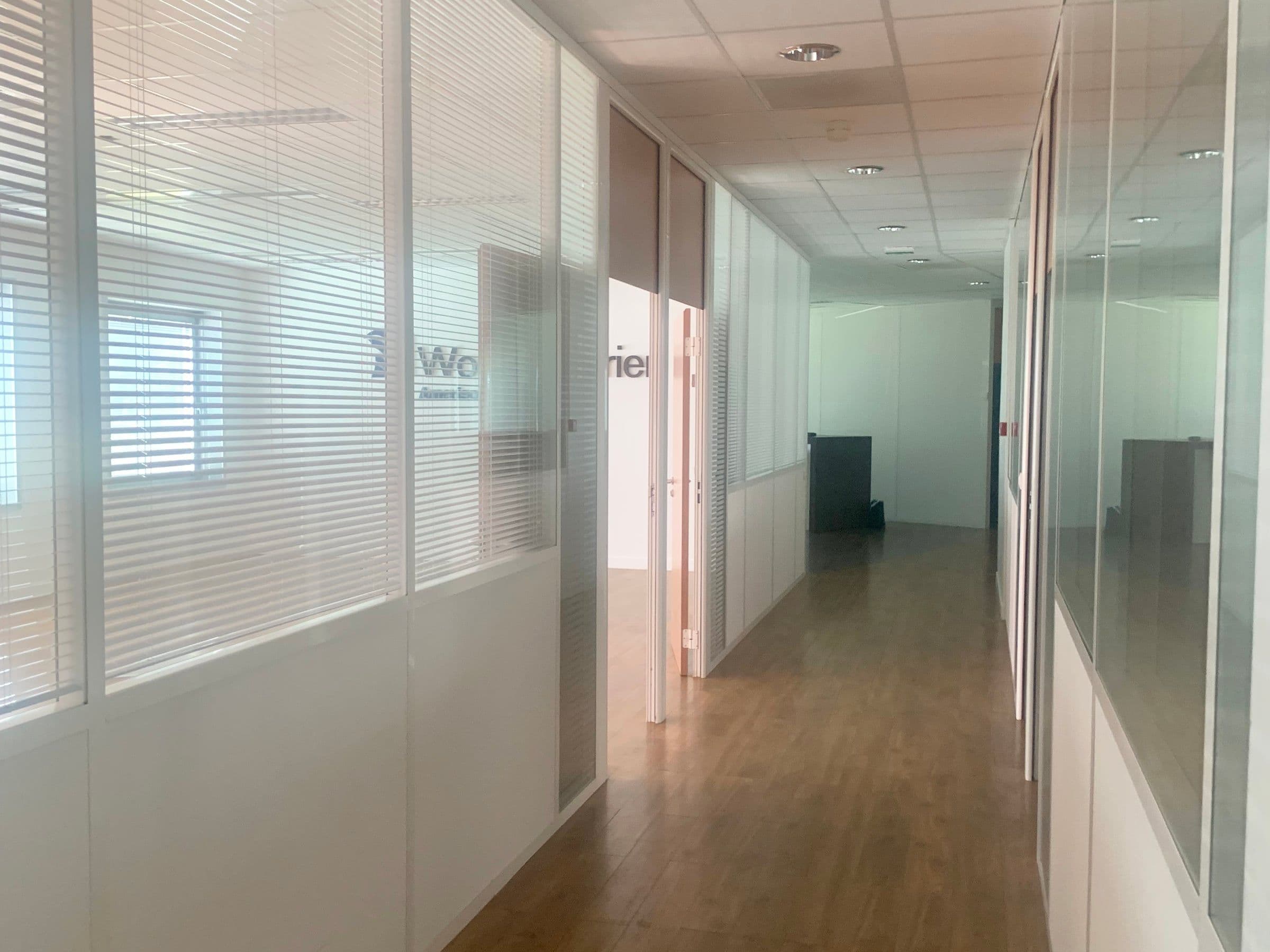 Avant travaux : couloir de bureaux aux cloisons vitrées à stores intégrés, sol en parquet stratifié et faux plafond à dalles acoustiques.