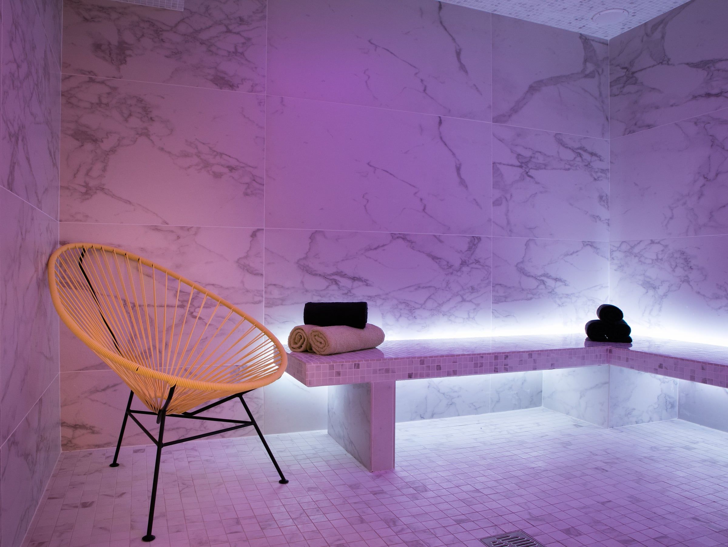 Hammam de spa habillé de marbre, éclairage d'ambiance rose, banc en mosaïque et fauteuil suspendu en cordage orange.