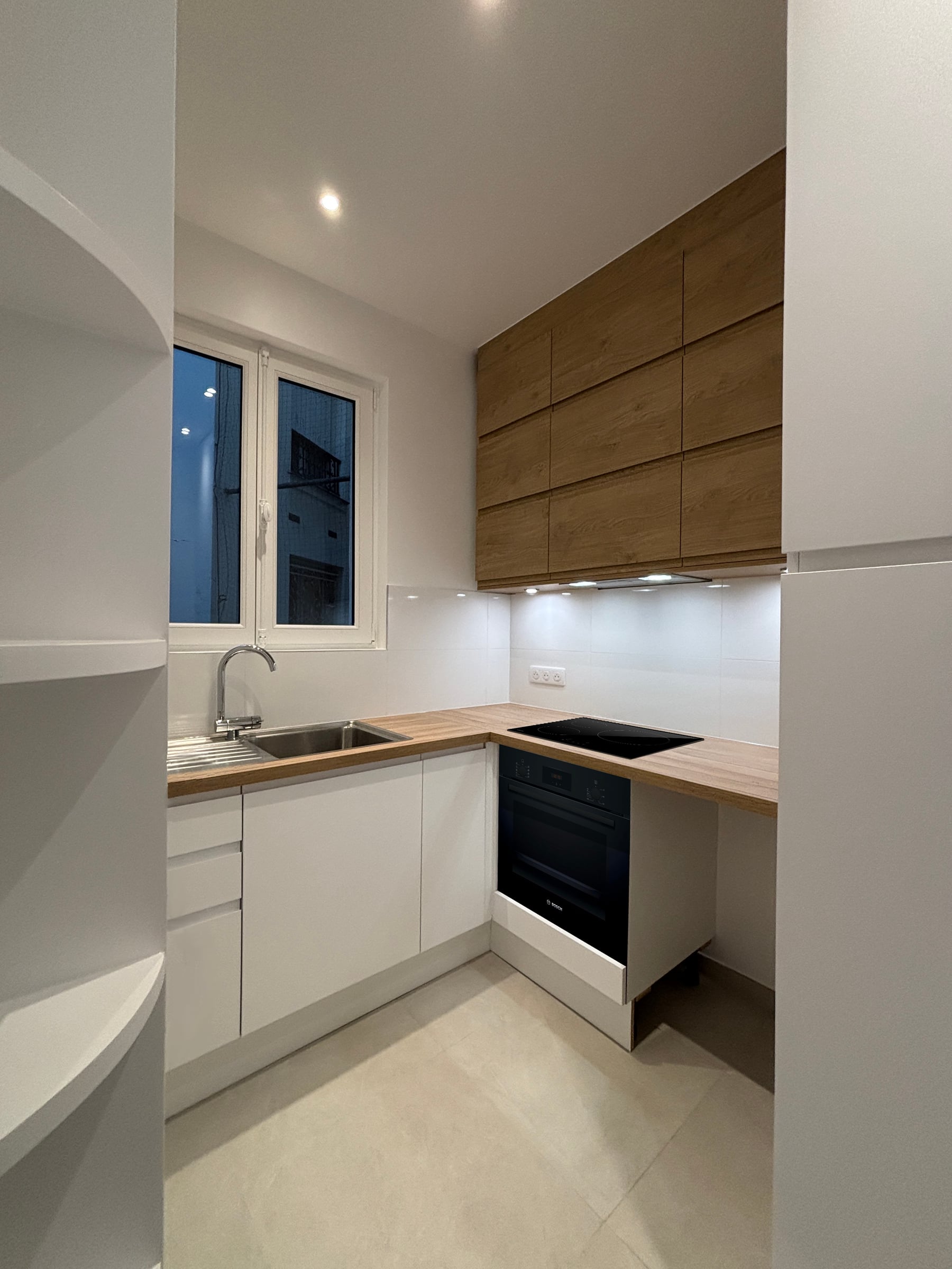 Cuisine contemporaine en U à façades blanches, plan de travail en chêne et four encastré, rénovation de cuisine éclairée par une fenêtre.
