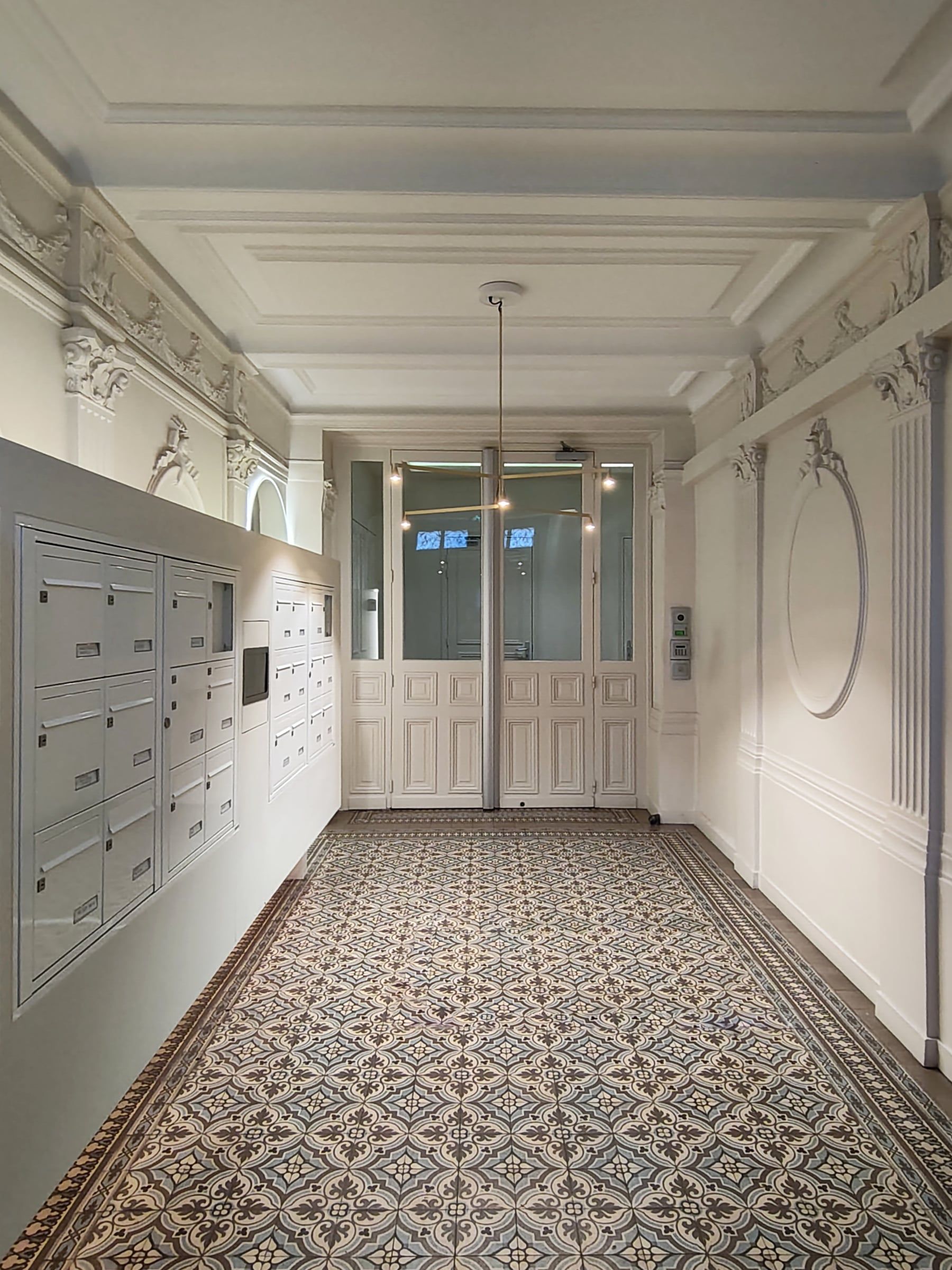 Rénovation de parties communes : hall haussmannien à porte vitrée, sol en carreaux de ciment, corniche moulurée et chapiteaux corinthiens en plâtre.