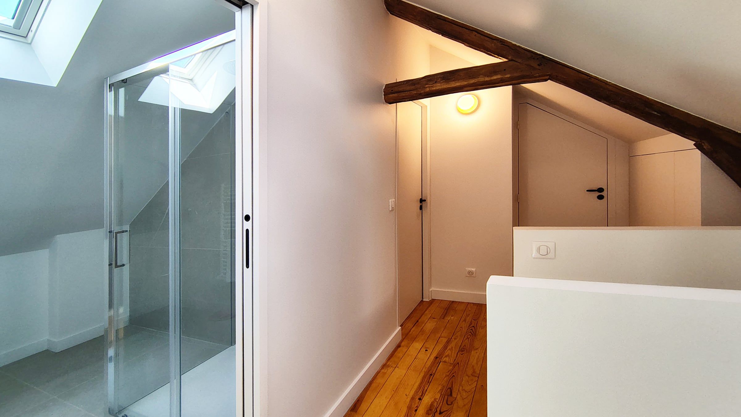 Palier sous combles rénové par un architecte d’intérieur, associant parquet bois, poutres anciennes et salle de bain vitrée sous fenêtre de toit.