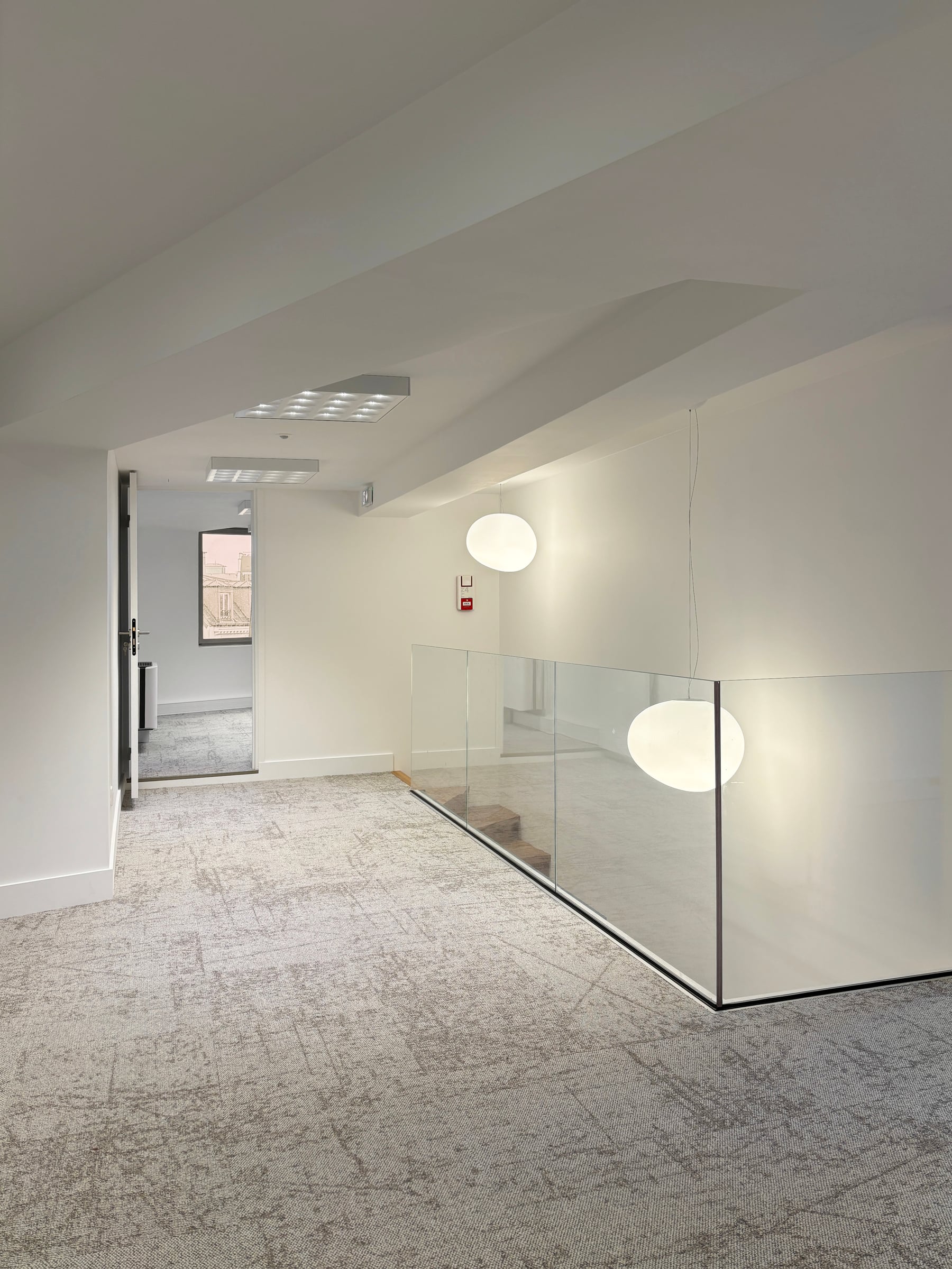 Palier de bureaux avec garde-corps en verre, suspensions globe en verre opalin et moquette grise texturée, aménagement de bureaux contemporain