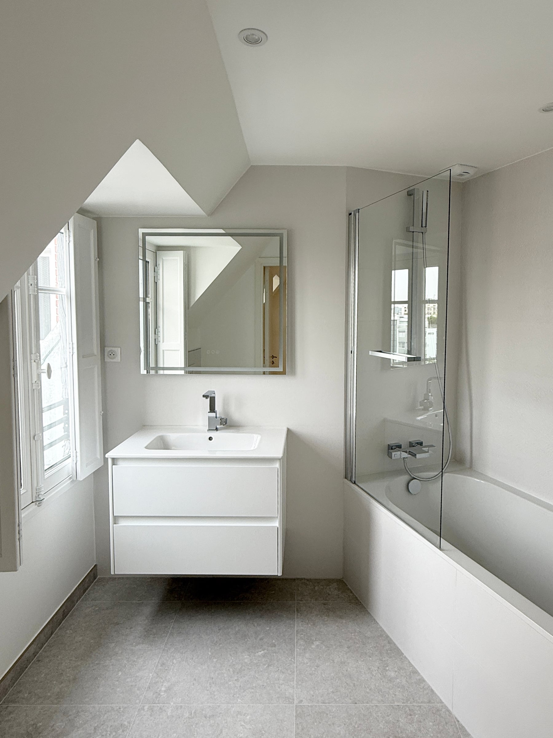 Salle de bains sous rampant : meuble vasque suspendu blanc, miroir cadré, baignoire, pare-douche vitré et sol en grès gris, aménagement de salle de bain.