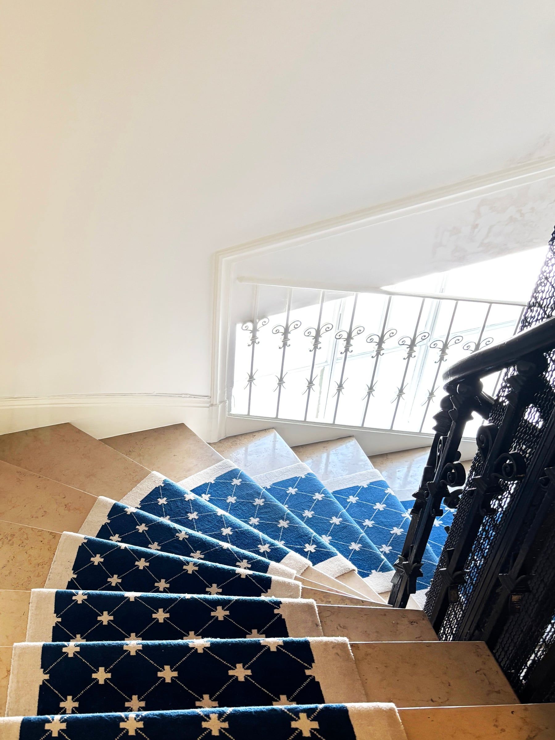 Escalier tournant en pierre claire avec tapis bleu bordé d’écru, garde-corps en fer forgé et fenêtre protégée par une grille.