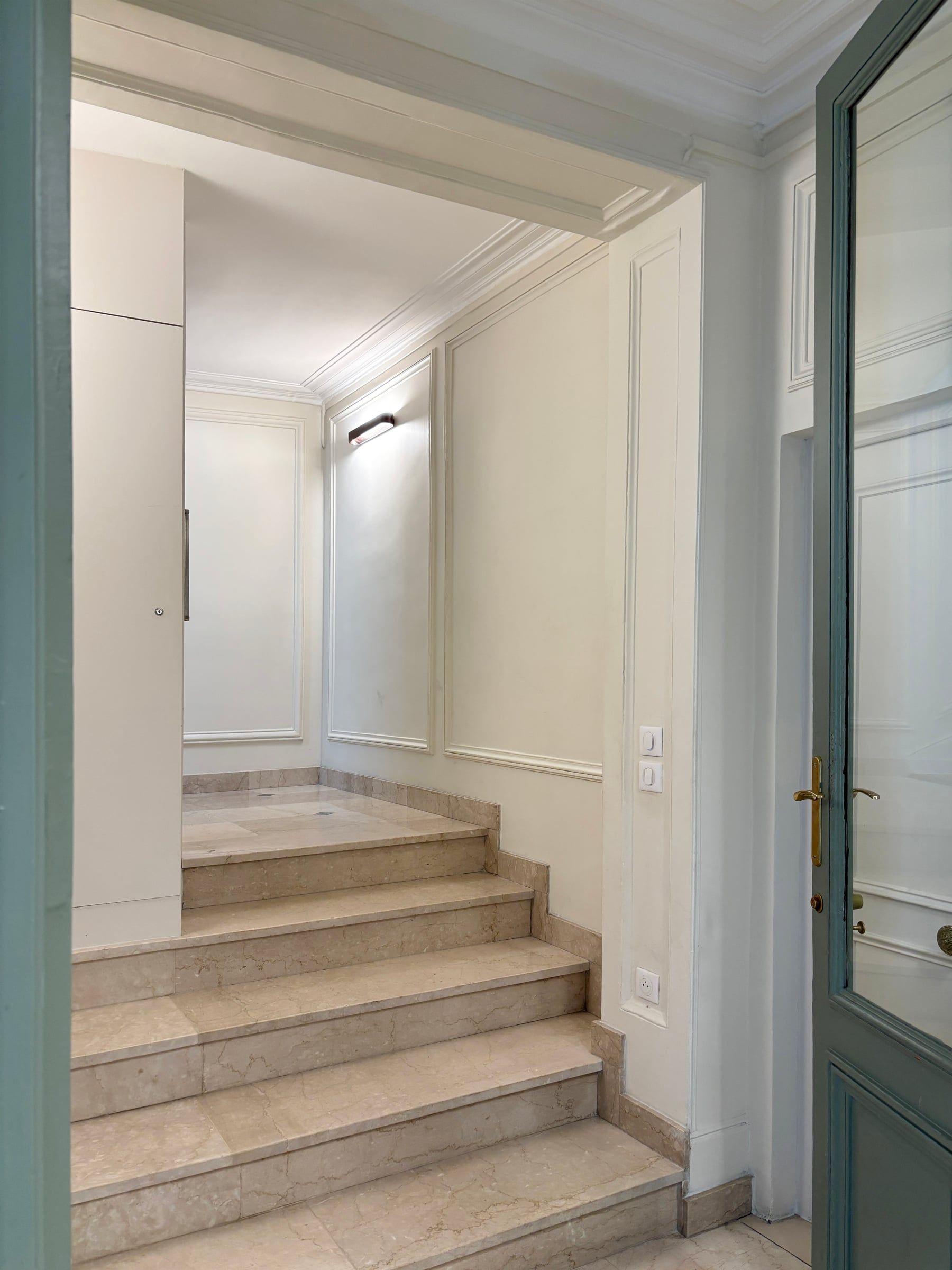 Demi-palier haussmannien avec emmarchement en pierre menant à un repos, boiseries et moulures crème, porte vitrée à imposte en menuiserie vert d'eau, en copropriété.