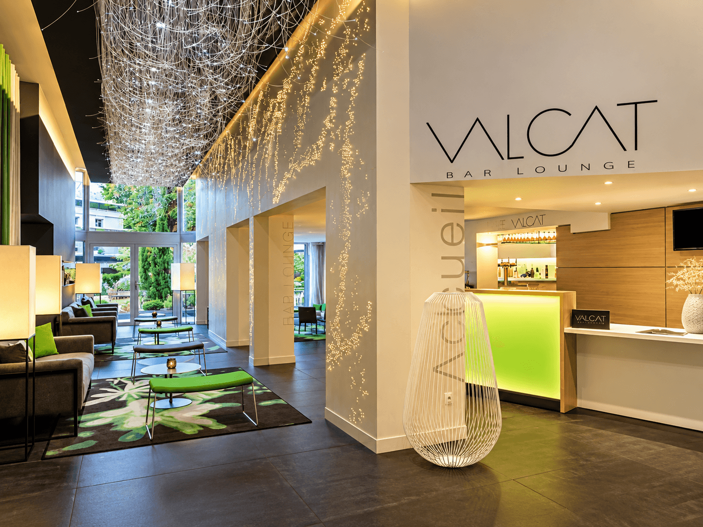 Hall d'entrée d'hôtel avec enseigne murale Valcat, suspension de fibres optiques, lampe sculpturale en fil et comptoir rétroéclairé vert.