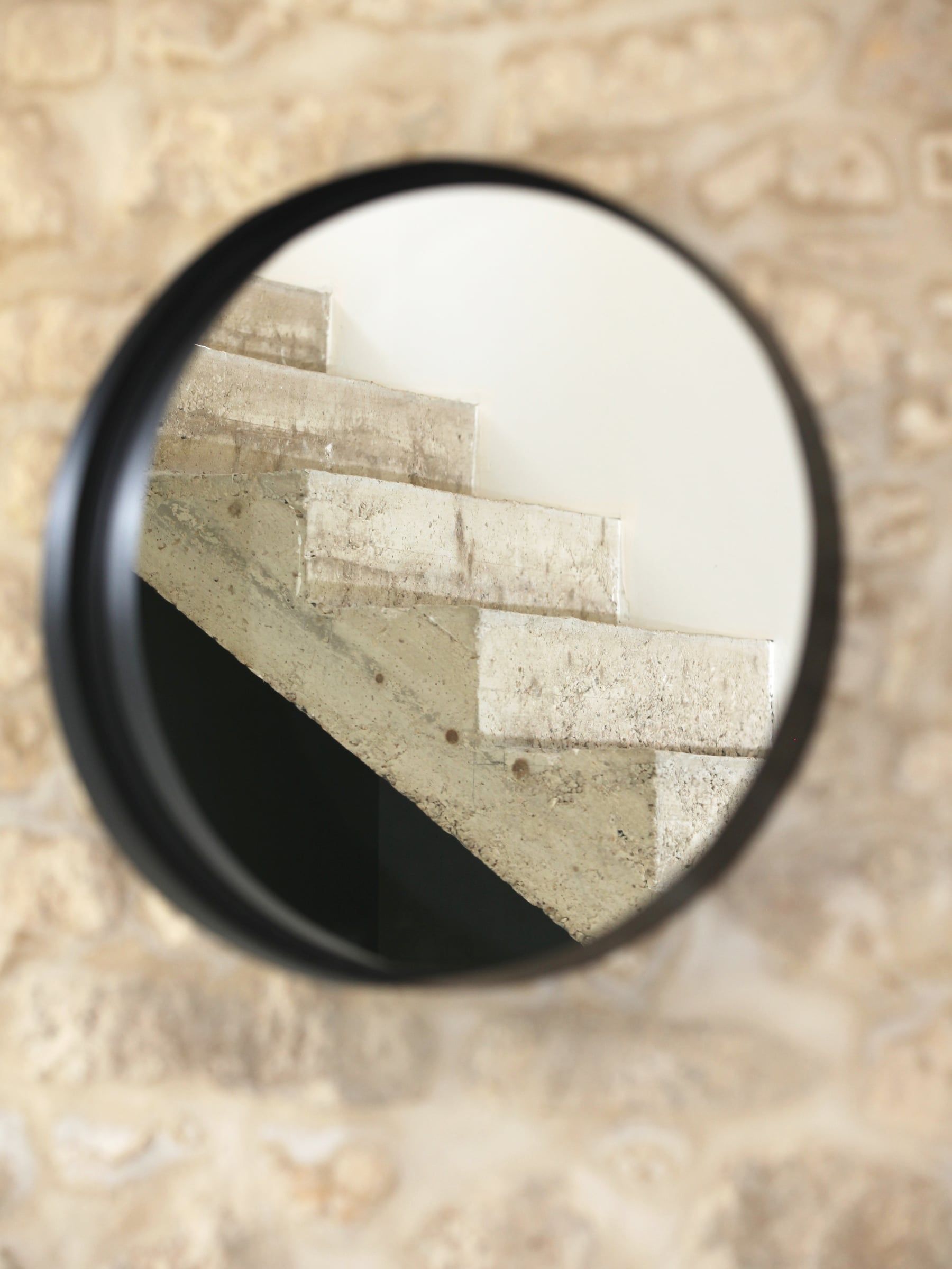 Détail de l’escalier en pierre révélé dans un miroir rond à cadre noir, posé sur un mur de moellons de la maison rénovée.