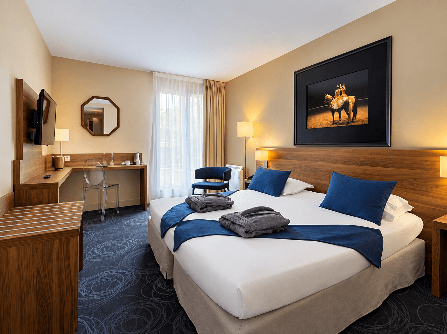 Chambre d'hôtel avec lit double, tête de lit et bureau en noyer, coussins bleus et tableau équestre encadré au-dessus du lit.