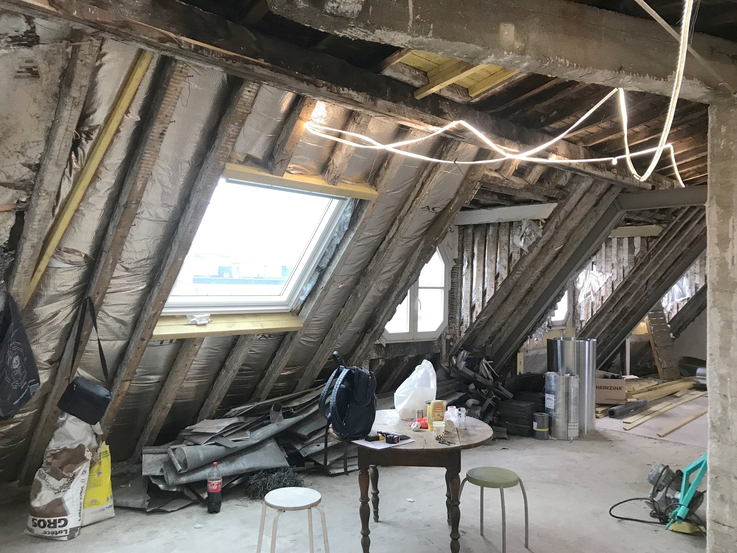 Avant travaux : combles bruts à charpente apparente, isolant en sous-toiture, fenêtres de toit existantes et sol béton encombré de gravats.