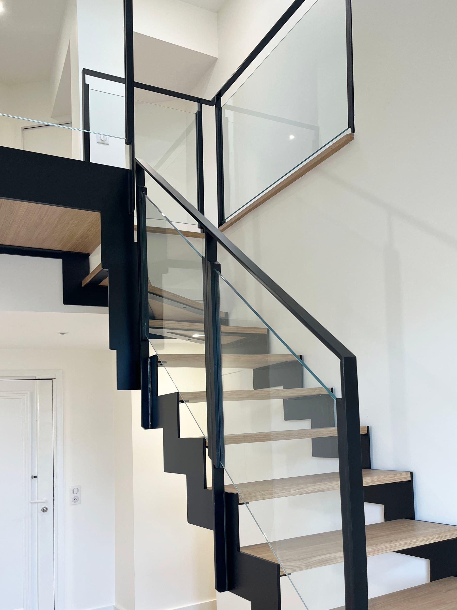 Escalier à limon crémaillère en acier noir, marches en chêne clair et garde-corps vitré desservant une mezzanine, agencement sur mesure.