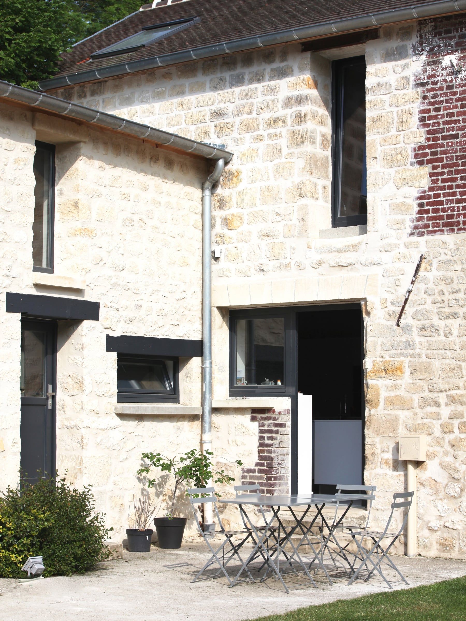 Réhabilitation d’une façade secondaire en pierre et brique, avec fenêtres à cadres noirs, gouttière métallique et petite terrasse de jardin.