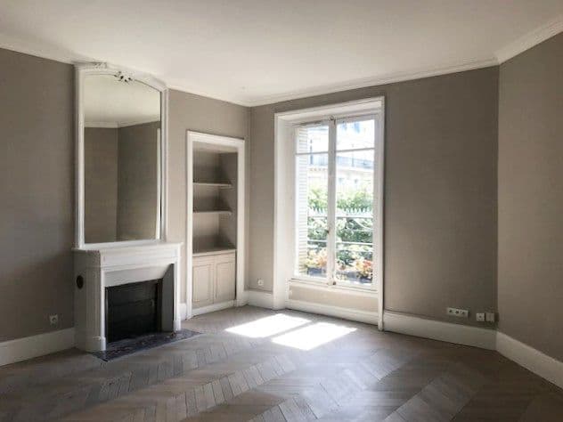 Avant travaux : salon haussmannien aux murs taupe, cheminée en marbre surmontée d'un trumeau, niche à étagères et parquet en chevrons à rénover.