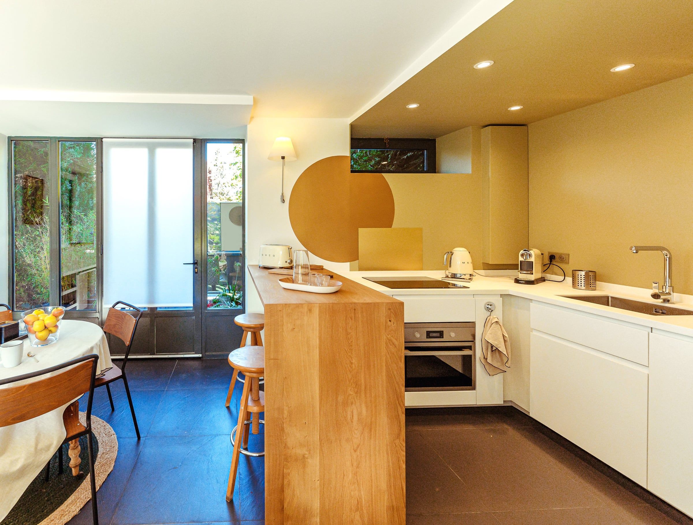 Cuisine ouverte aux façades blanches et îlot en chêne massif, mur ocre ponctué d'un disque peint et baie vitrée sur jardin, rénovation de cuisine.