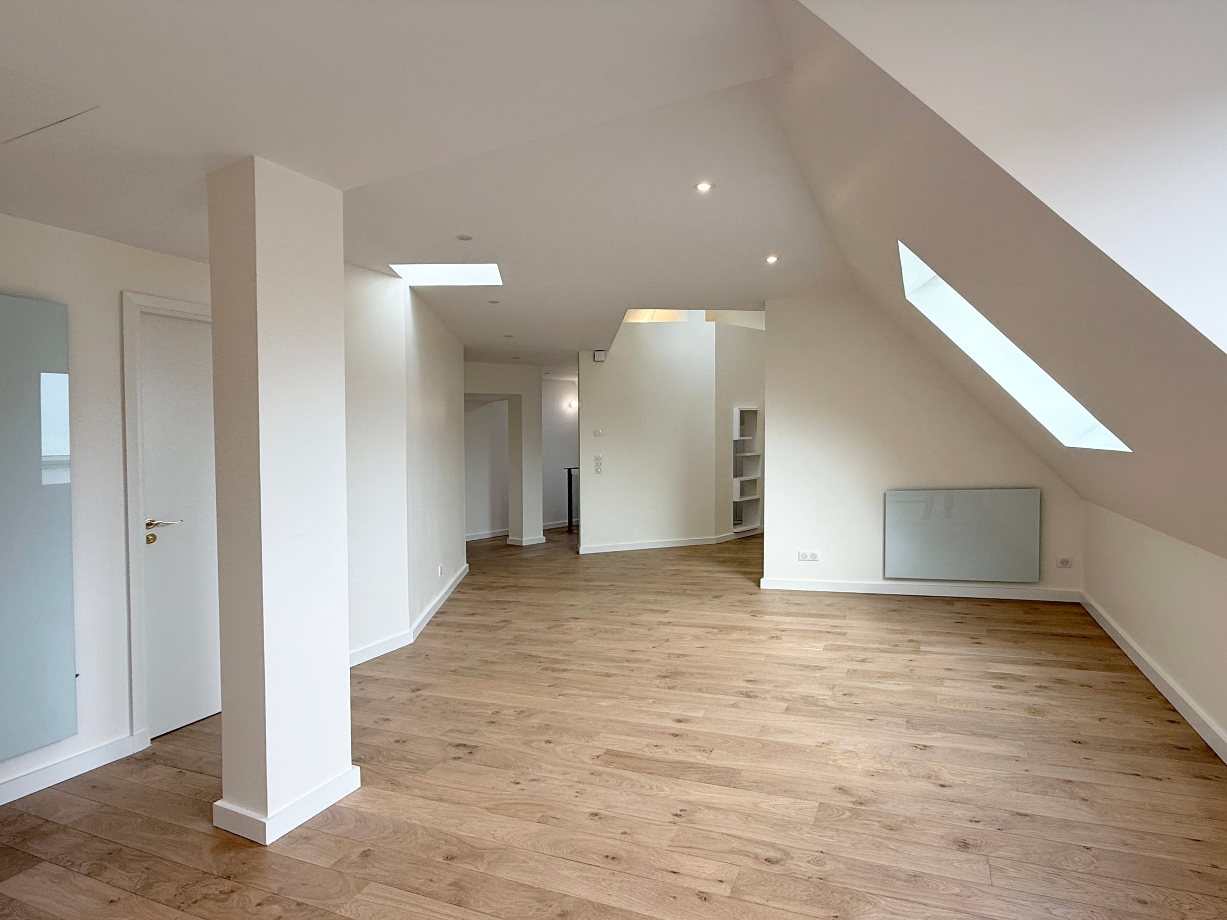 Pièce de vie sous combles avec parquet en chêne clair, poteau structurel blanc, fenêtre de toit en pente et niches d'étagères encastrées.