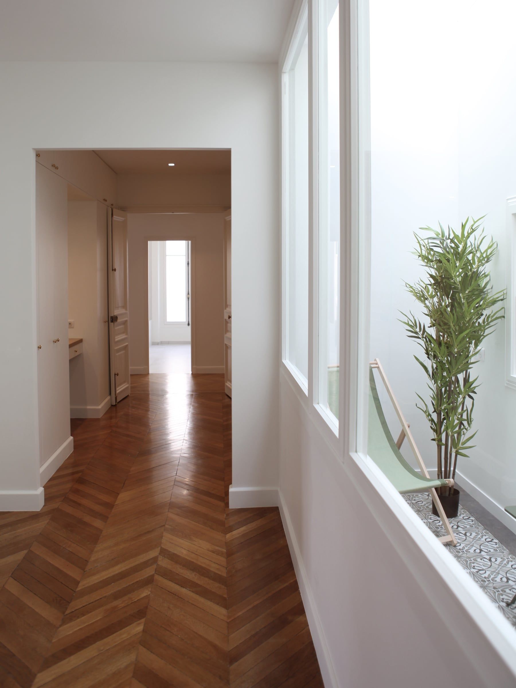 Couloir haussmannien au parquet en chevrons longeant une verrière vitrée ouvrant sur un patio, en enfilade de pièces blanches après rénovation d'appartement.