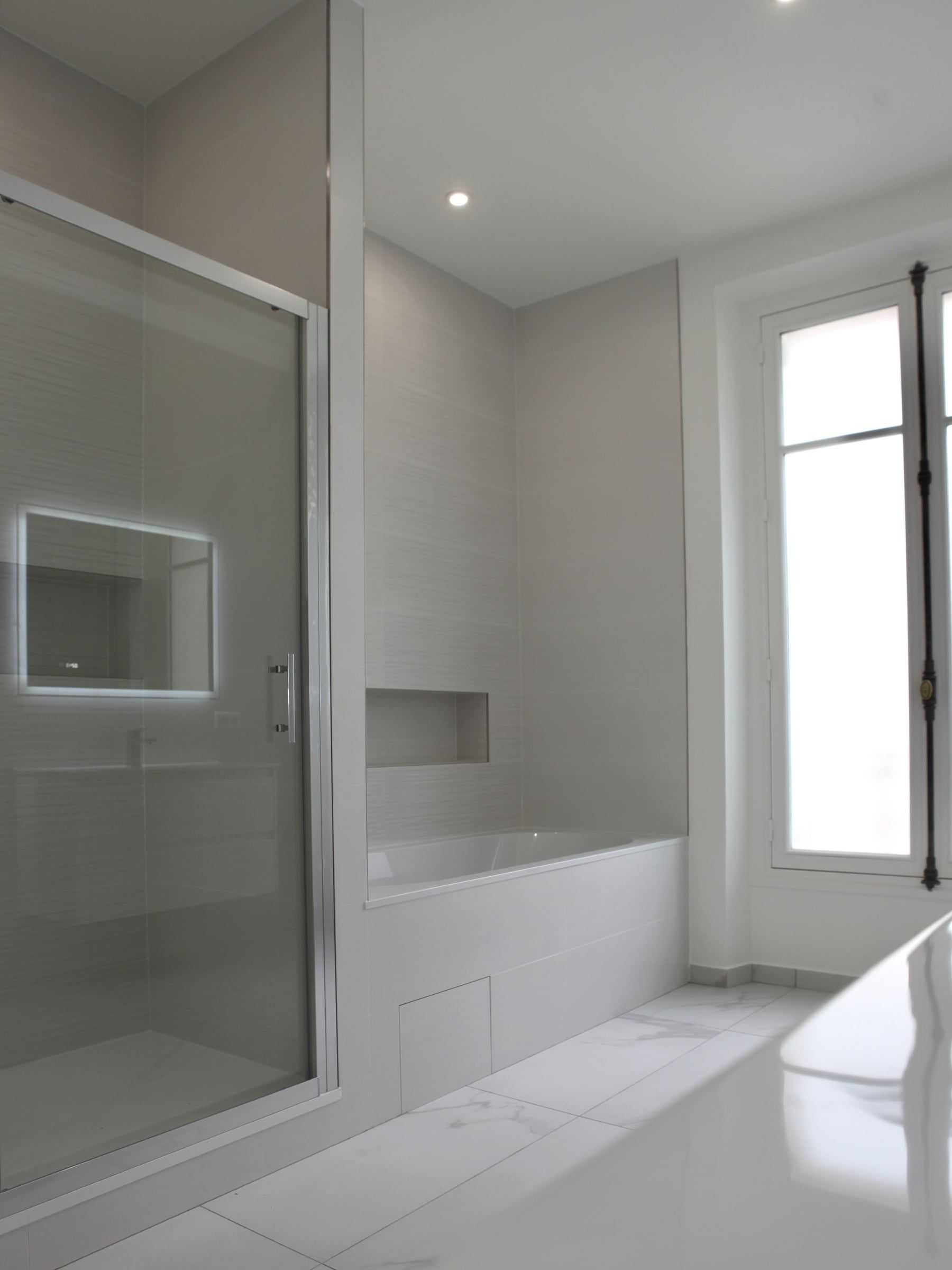 Salle de bain au carrelage gris et marbre blanc, baignoire encastrée, cabine de douche vitrée et fenêtre à croisillons après aménagement de salle de bain.