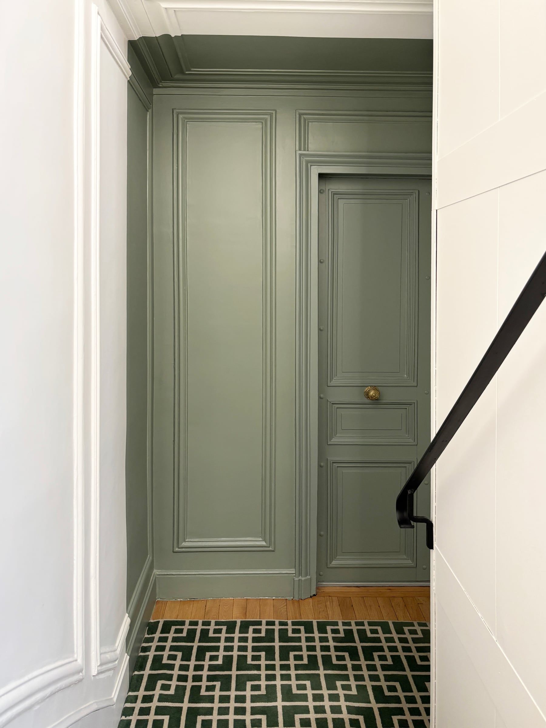 Couloir haussmannien aux boiseries vert sauge, porte à moulures et poignée en laiton, parquet et tapis vert géométrique, rampe en fer noir, rénovation de parties communes.