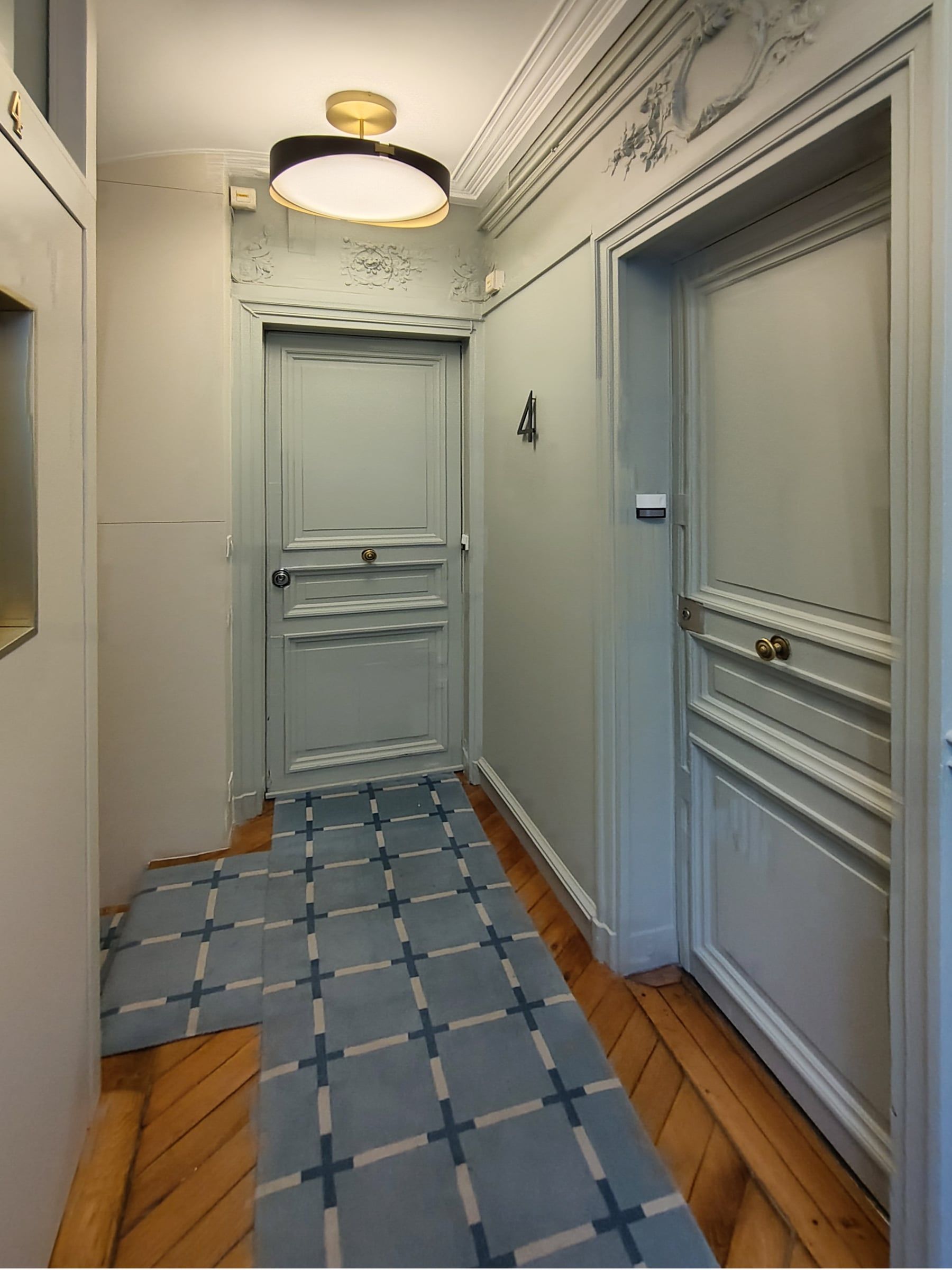 Palier d'étage à porte palière grise moulurée, stucs en imposte, tapis bleu à motif quadrillé et plafonnier en laiton et opaline.