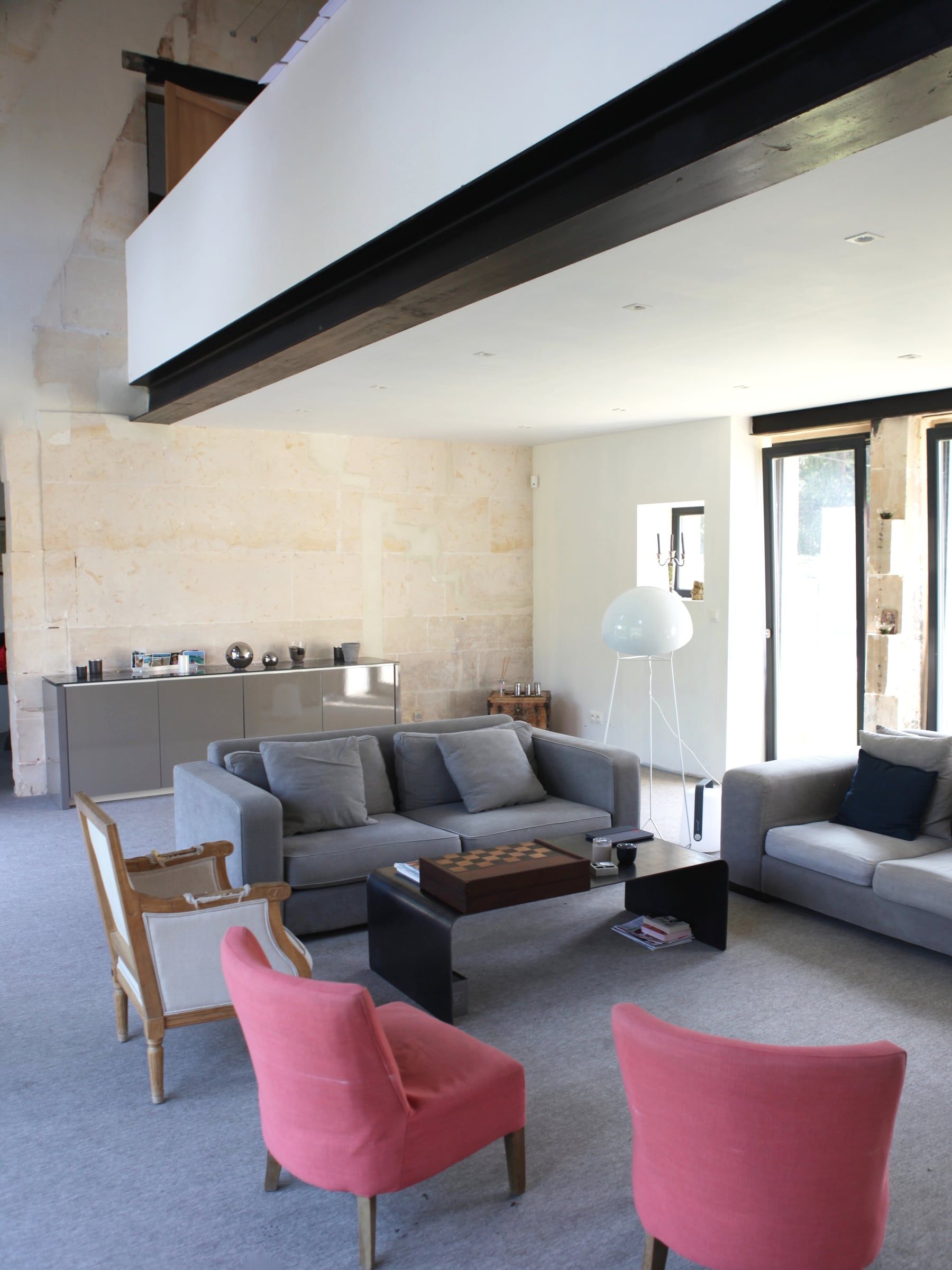Aménagement du salon sous mezzanine, avec mur en pierre apparente, poutre sombre, mobilier gris et grandes portes vitrées vers le jardin.