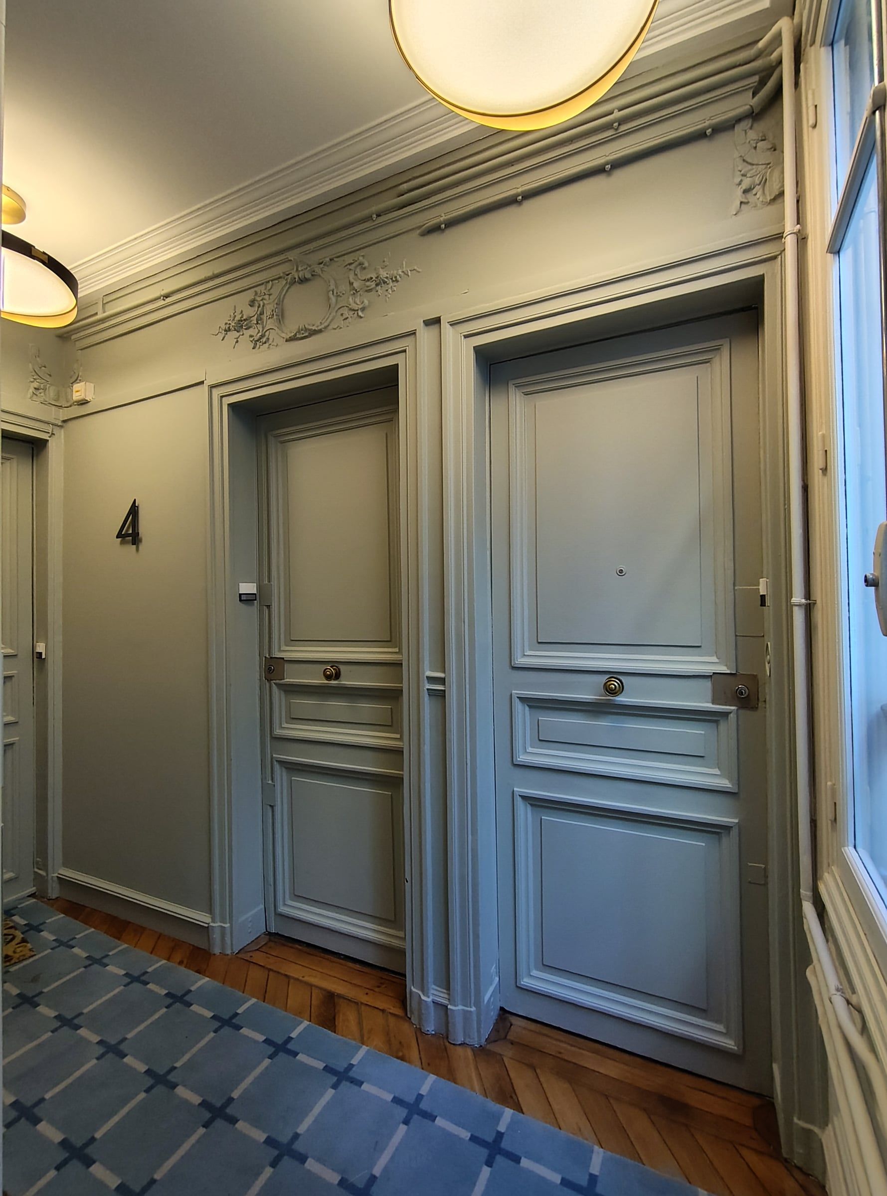 Hall d'immeuble : palier desservant deux portes grises moulurées, parquet en point de Hongrie, tapis bleu quadrillé, stucs en imposte et plafonnier laiton.