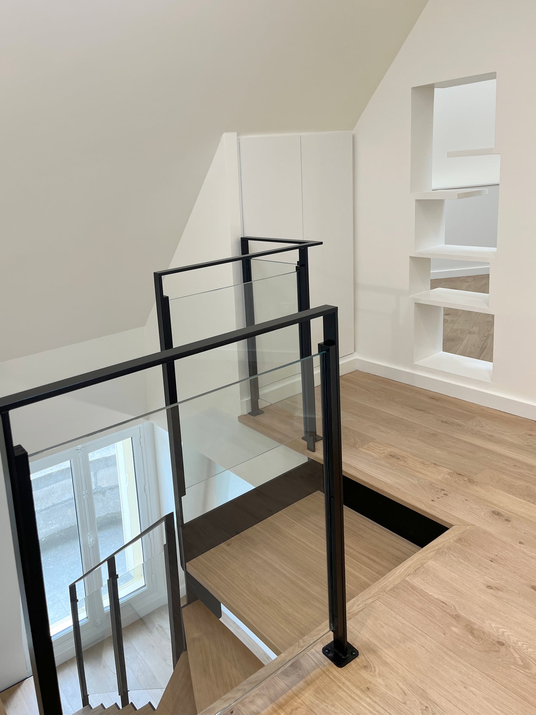 Palier ouvert sur l'escalier, garde-corps en métal noir et verre, parquet en chêne clair et niches blanches encastrées sous rampant sur mesure.