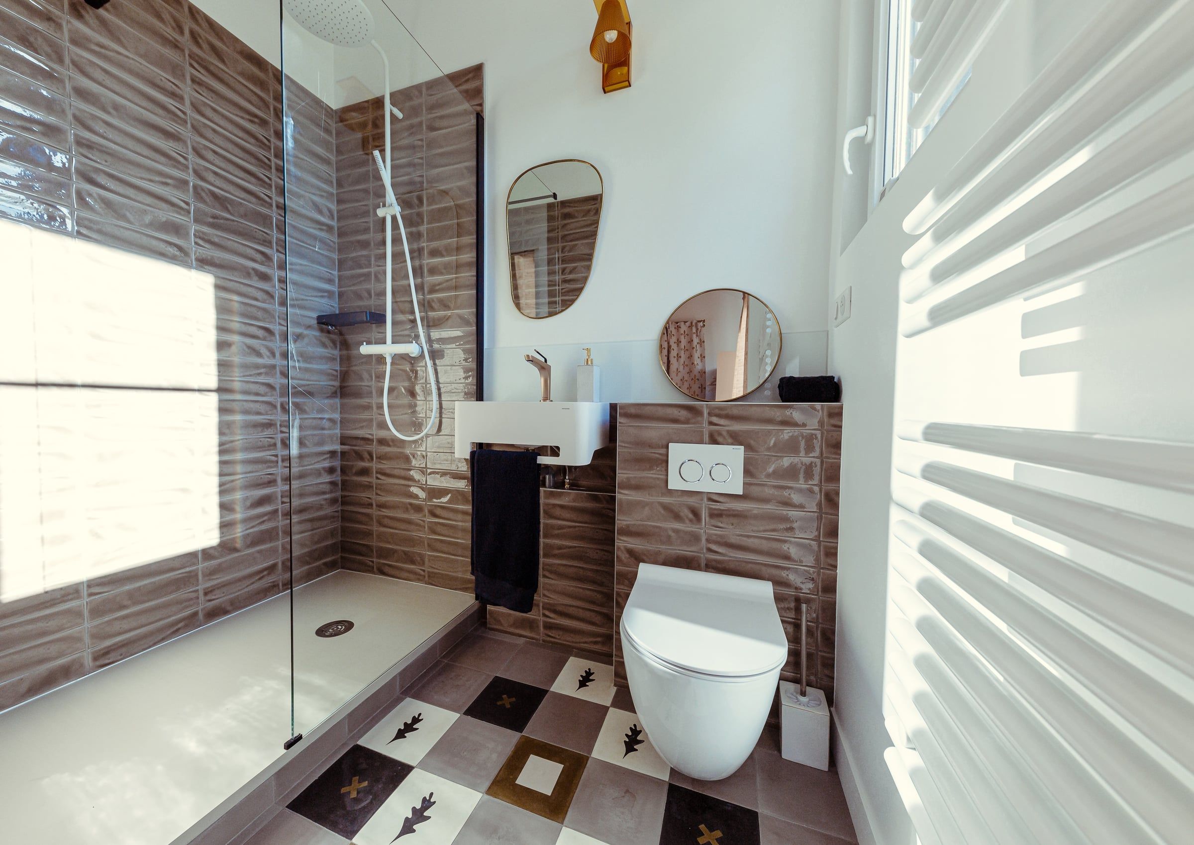 Salle de bain avec douche à l'italienne, faïence relief brun, sol en tomettes à motifs et vasque suspendue, aménagement de salle de bain en lumière du jour.